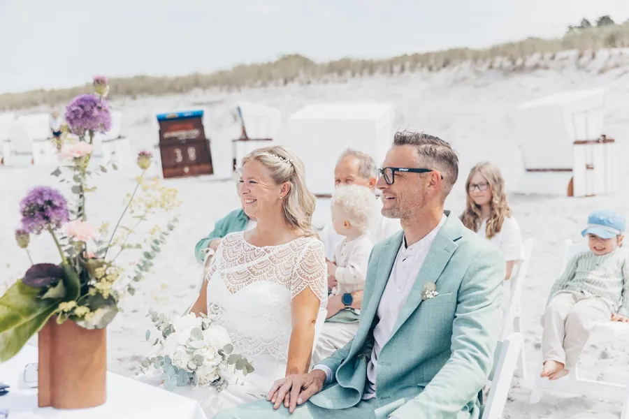 Brautpaar sitzt lächelnd bei der Strandhochzeit am Ostseestrand von Ahrenshoop, dahinter Gäste und Strandkörbe.