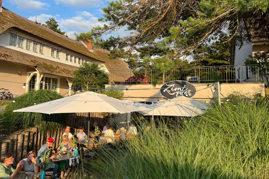 Außenterrasse des Restaurants ZIMTZIEGE im STRANDHAUS Ahrenshoop mit Gästen, Sonnenschirmen und Reetdachhaus im Abendsonnenlicht.