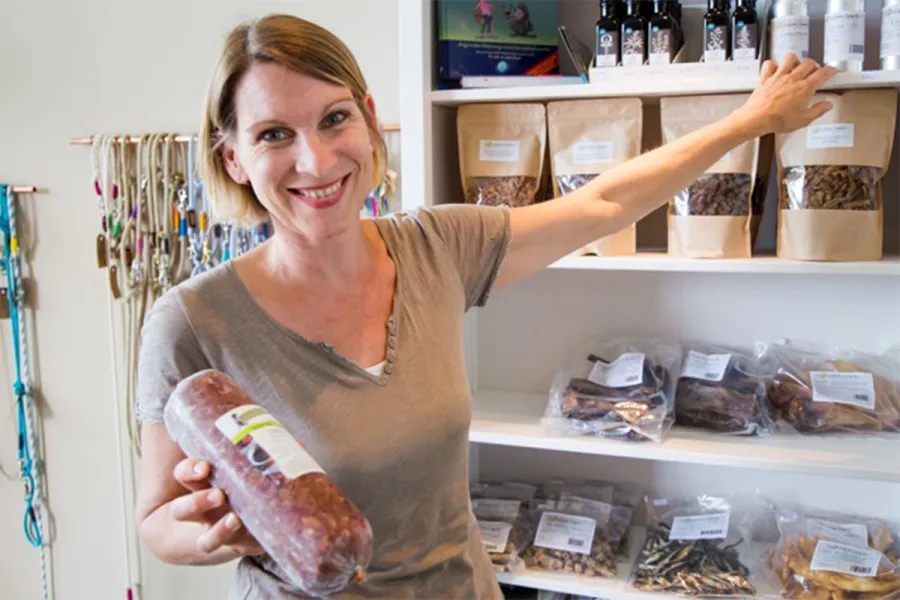 Lächelnde Mitarbeiterin hält eine Rolle Hundefutter vor einem Regal mit Futter und Leinen im Hundeshop URLAUBSFUTTER.