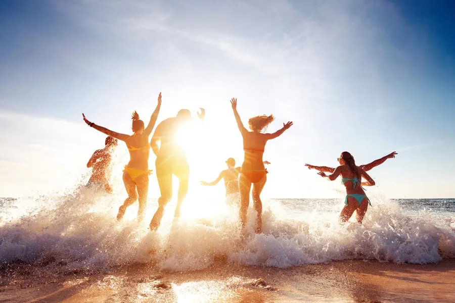 Gruppe jubelnder Menschen springt im Gegenlicht durch Ostseewellen am Strand bei strahlend blauem Sommerhimmel.