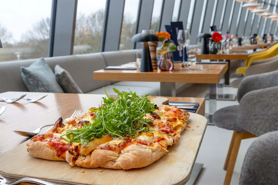 Rechteckige Pizza mit frischem Rucola auf einem Holzbrett vor der Fensterfront eines Restaurants mit Meerblick.