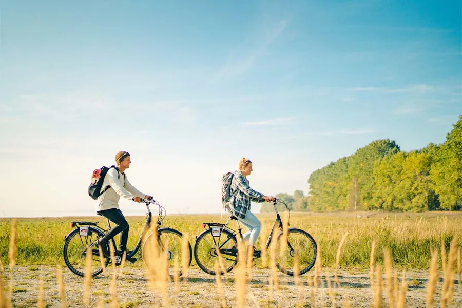 Zwei Radfahrer mit Rucksaecken fahren durch eine sonnige Landschaft auf dem Fischland-Darß an der Ostsee.