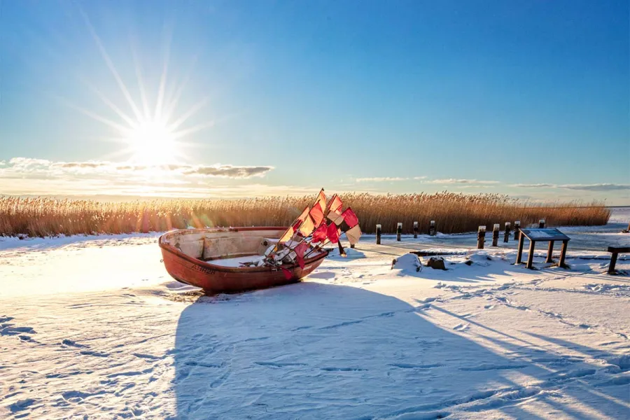 Verschneiter Hafen in Ahrenshoop mit altem Fischerboot und Reusenpfählen im Gegenlicht der Wintersonne.