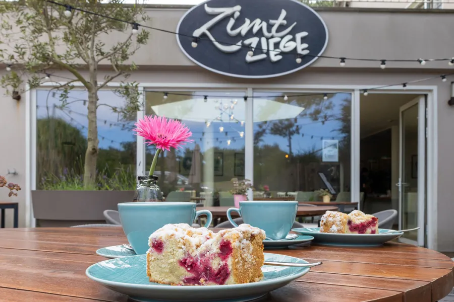 Zwei Stück Streuselkuchen mit Beeren und zwei türkise Tassen auf einem Holztisch vor dem Restaurant ZIMTZIEGE in Ahrenshoop.
