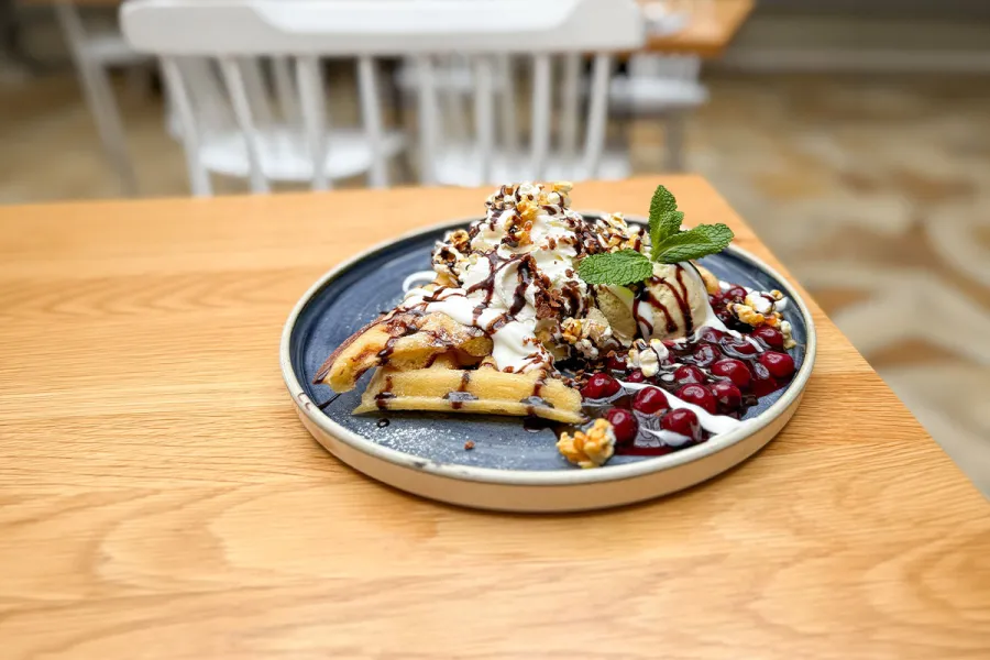 Blauer Teller mit einer belgischen Waffel, Sahne, Schokosauce, Eis und heissen Kirschen auf einem Holztisch.