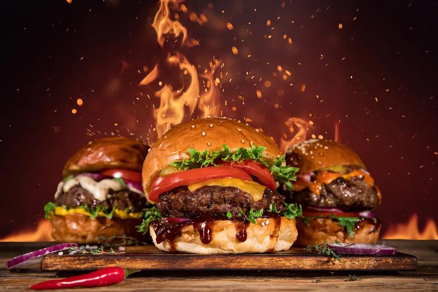 Drei Burger mit Rindfleisch, Käse und Tomate auf einem Holzbrett vor lodernden Flammen, davor eine rote Chilischote.