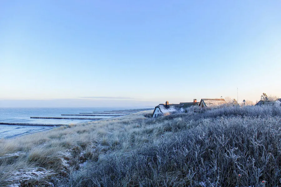 Reifbedeckte Dünenlandschaft mit Reetdachhäusern am Ostseestrand auf dem Fischland-Darß an einem klaren Wintertag.