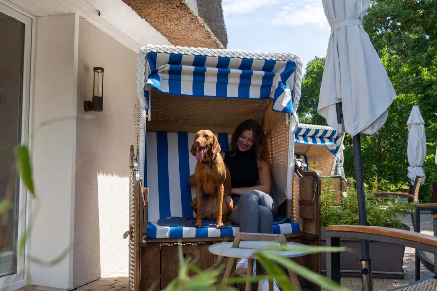 Frau sitzt mit einem hellbraunen Hund in einem blau-weißen Strandkorb auf der Terrasse des STRANDHAUS Ahrenshoop.