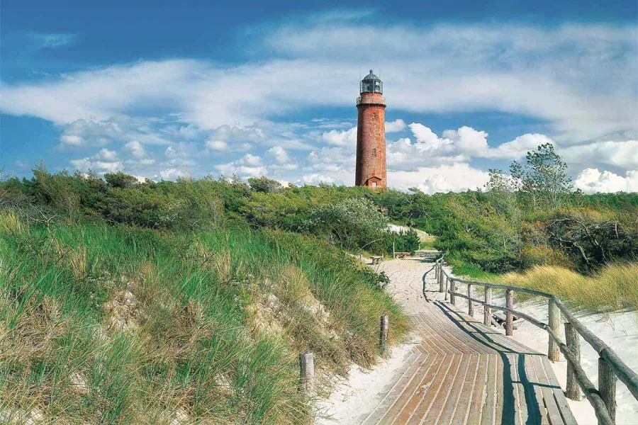 Leuchtturm Darß hinter grünen Dünen mit Holzsteg auf dem Fischland-Darß an einem sonnigen Sommertag.