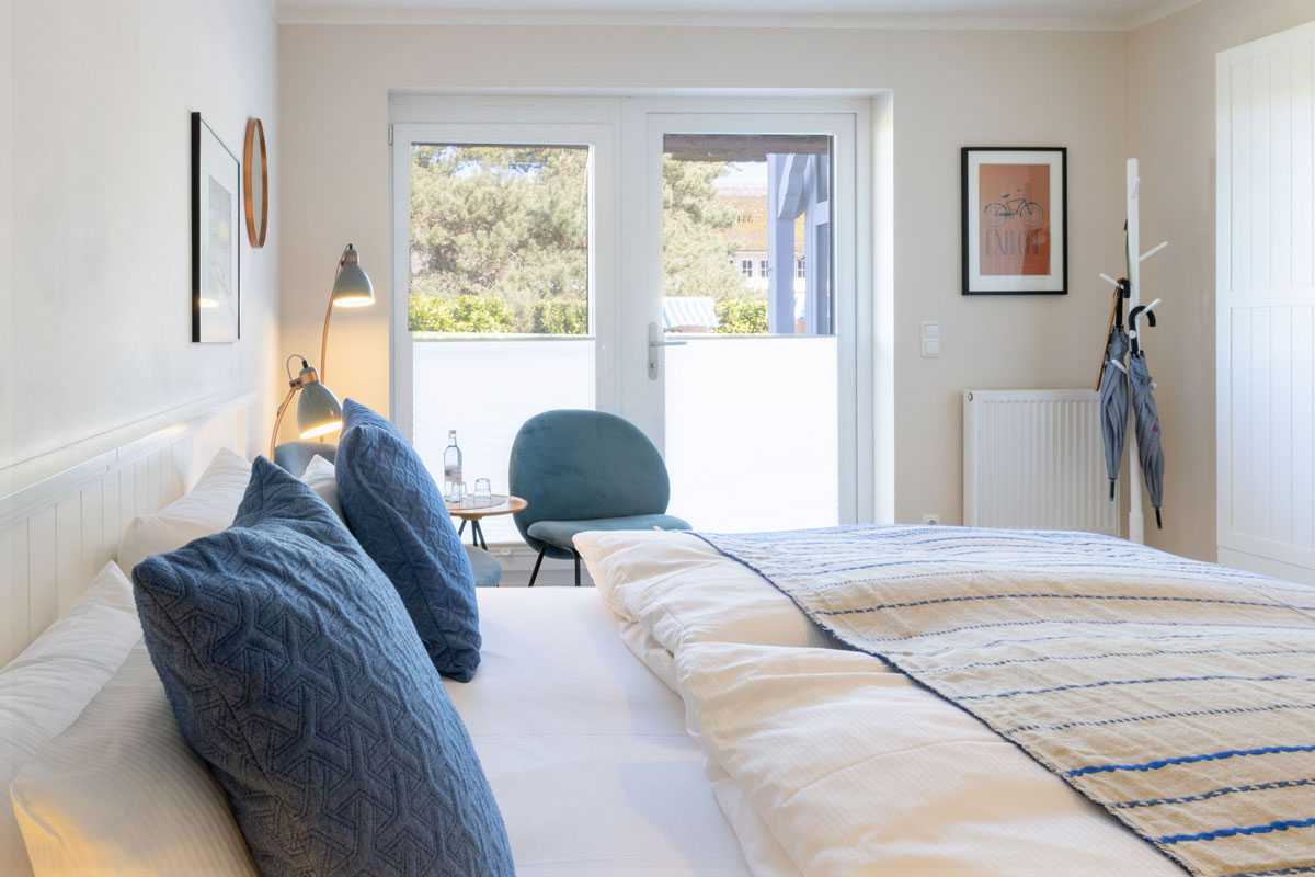 Doppelzimmer Petit im STRANDHAUS Ahrenshoop mit Doppelbett, blauen Kissen und Terrassentür zum Grünen.