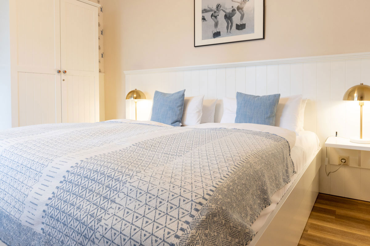 Doppelbett im Doppelzimmer Petit des STRANDHAUS Ahrenshoop mit blauer Tagesdecke, goldenen Lampen und Einbauschrank.