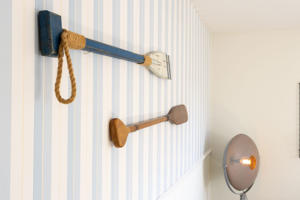 Maritimes Wanddekor im Apartment des STRANDHAUS Ahrenshoop mit zwei Holzrudern vor blau-weißer Streifentapete.