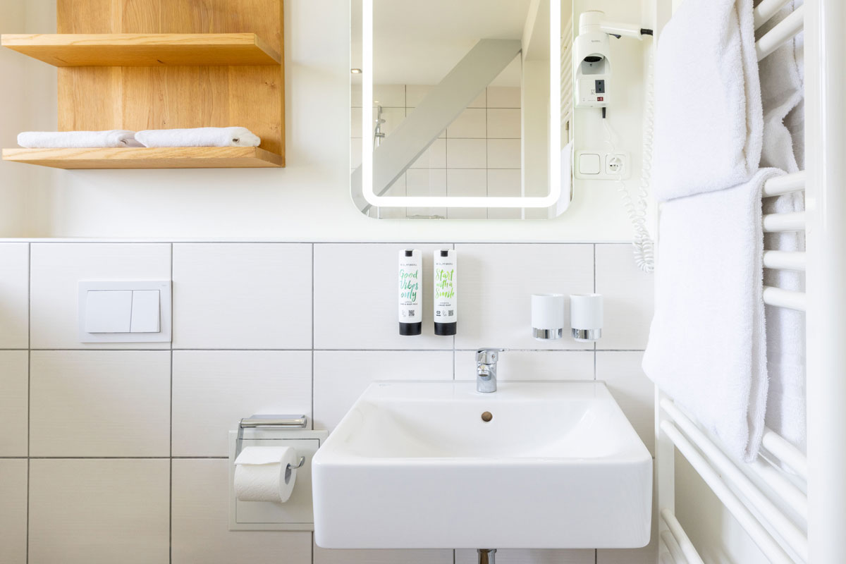 Bad der Familiensuite im STRANDHAUS Ahrenshoop mit Waschbecken, beleuchtetem Spiegel, Holzregal und Handtuchhalter.