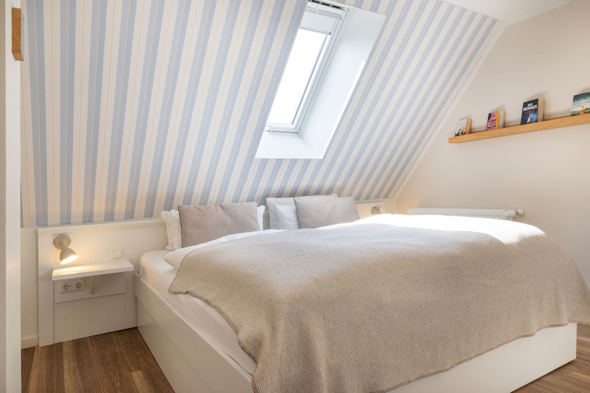 Doppelbett im Familienzimmer des STRANDHAUS Ahrenshoop mit blau-weißer Dachschräge, Wandlampen und Bücherregal.